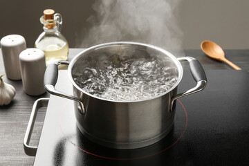 Cooking pot with boiling water and stove on wooden table against grey background, closeup