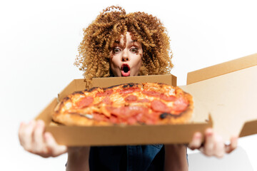 Excited woman with curly hair holding a delicious pepperoni pizza against a bright white background, expressing surprise and joy in a casual, trendy outfit