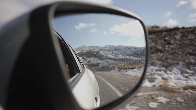 A passenger records a friend in the rearview mirror. An attractive stylish girl with a tattoo rides in a car and enjoys a trip, leaning out of the backseat window and catching gusts of wind with her