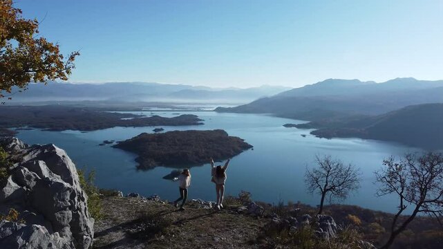 Two young woman friends stand at the top of a mountain, hugging and celebrating their achievement with joy and laughter. Behind them stretches a breathtaking view of an endless valley dotted with