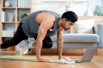 Obraz premium Young asian man doing home workout while watching online tutorial on laptop computer in living room at home, determined and motivation for health, healthy lifestyle and body strength for exercise.