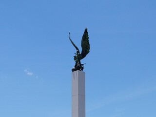 A bronze eagle sculpture with wings spread sits atop a tall modern white pillar, dramatically contrasting with the clear blue sky in a coastal setting.