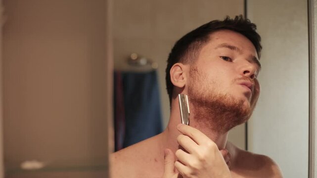 A man carefully trims his beard with a straight razor, focused on his reflection in the bathroom mirror. It's a quiet moment of precision and ritual in his morning routine.