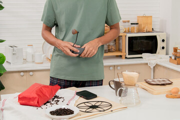 Middle-aged bearded Asian man grinds coffee beans with traditional coffee grinder using hand-crank to make the coffee beans powdered for drink in morning at home. Healthy lifestyle concept.