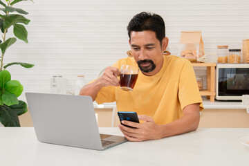 Middle-aged bearded Asian man holding laptop walks into kitchen to grab some milk for breakfast. Freelancer working from home and reading news in morning. Male successful entrepreneur working at home.