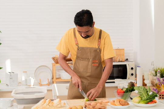 Asian man with beard sitting makes sandwich and divide two peice with knife in kitchen for breakfast in morning. Male wear apron enjoy make food for health care in house, healthy food at home.