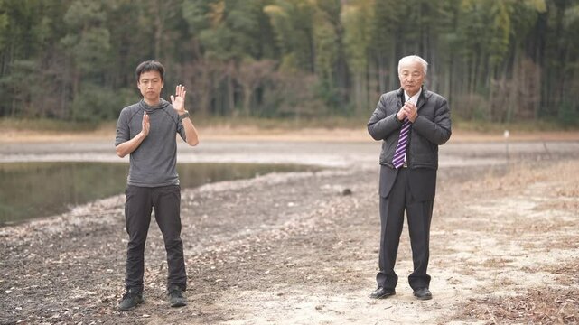 There is a lot of greenery in the bamboo forest pond at noon in spring in Japan. Japanese men in the late 70s stand in suits. He and his son, in his 30s, are practicing by watching Tai Chi videos.