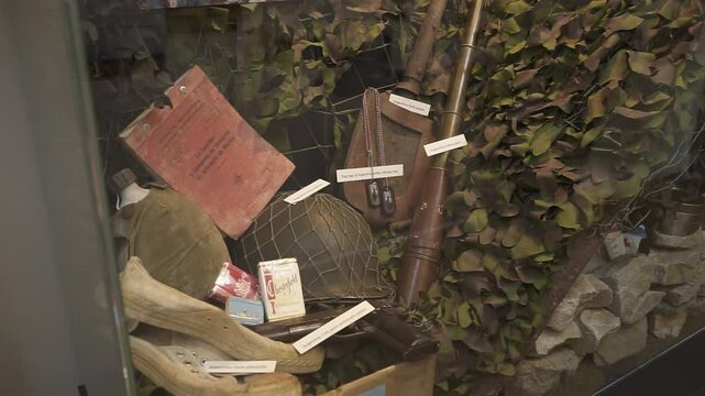 Military artifacts from world war ii, including a helmet, canteen, rifle, documents, cigarettes, and camouflage netting, are showcased in a museum display case