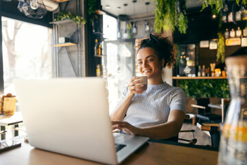 Portrait of young interracial freelancer sitting in coffee shop, drinking coffee and typing on...