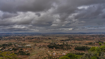 Panorama of the countryside, view from a height. Village houses, cultivated fields, trees, unpaved red-soil roads. Mountains on the horizon. Picturesque dark clouds in the sky. Madagascar.