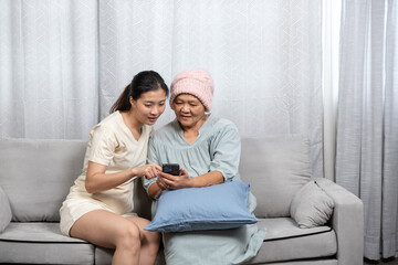 Young Asian woman comforting middle-aged Asian female cancer patient wearing soft pink knitted hat sitting together on cozy sofa. Warm emotional support, love, bonding, caring, healing, hope.