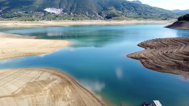 Drone flight over the dry Ramsko Jezero reservoir in Bosnia and Herzegovina, showing low water levels and exposed sandbanks, captured under the strong afternoon sun in October.