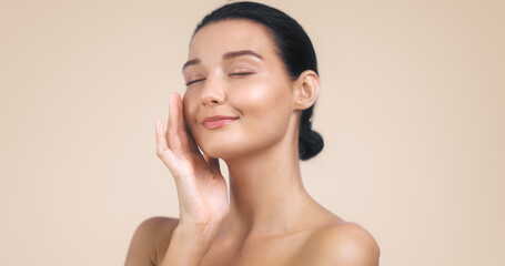 A beautiful woman with glowing skin and a serene smile, gently touching her face with closed eyes. The minimalist and soothing background highlights her natural beauty, making this image ideal for