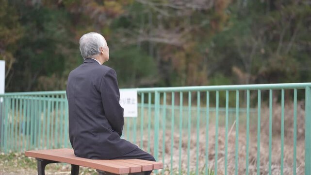 There is plenty of greenery in the spring noon park in Japan. A Japanese man from the late 70s was sitting on a bench in a suit.