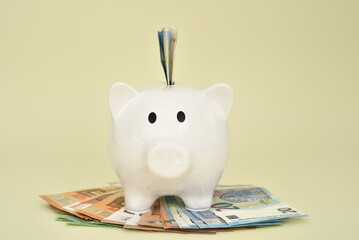 White piggy bank with banknote sticking out standing on the pile of different value euro banknotes on the pale yellow background. Saving money and financial concept. Copy space. Selective focus. 