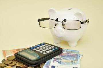 White piggy bank with glasses and calculator on the pile of coins and different value euro banknotes on the pale yellow background. Saving money and financial concept. Copy space. Selective focus. 
