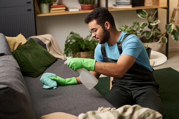 Man in uniform using detergent to clean sofa in the room during housework