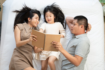 Obraz premium Top view of parent reading book with daughter on bed. happy family enjoy spending time together. Love bonding and insurance concept