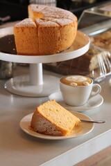 A slice of light, fluffy chiffon cake dusted with powdered sugar is presented on a plate next to a cup of latte 