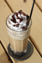 A tall glass of iced coffee with whipped cream, crumble chocolate cookie, and a straw sits on a wooden table.