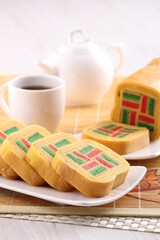traditional Indonesian layer cake, also known as Lapis Legit, with vibrant red and green checkered patterns, is sliced and presented on a white plate alongside a cup of tea and a teapot