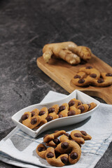 A serving of homemade ginger chocolate chip cookies, with their distinct paw-print shape, are beautifully arranged in a white dish on a striped napkin, with fresh ginger and more cookies on a wooden b
