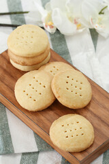 A small stack of round cookies with holes sits on a wooden board next to a white flower.