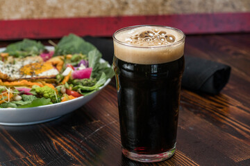 A glass of dark beer with a foamy head on it on table with salad