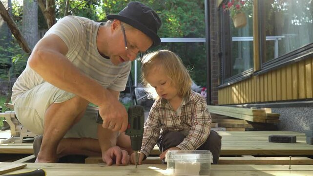 Father and his toddler work together on building a wooden terrace at home. The man uses a drill while the child observes and learns. Meaningful family moments. Give me a high five