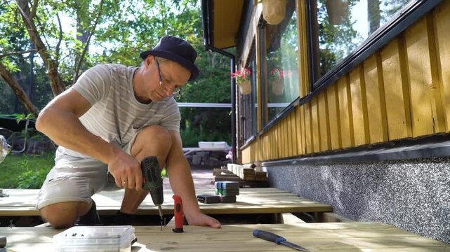 A man builds a wooden terrace in the backyard of a cozy house on a sunny summer day. He uses a drill and other tools while working on the decking. Surrounded by lush greenery and natural light.