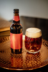 A frothy amber beer in a patterned glass sits beside a matching bottle with a red label on a hammered copper tray, with books and frames in background
