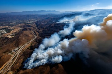 Fototapeta premium Aerial view depicts a wildfire spreading across a dry hillside environment, generating dense smoke plumes under a bright blue sky.