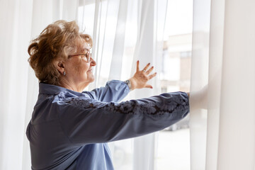 Elderly woman peers hopefully through sheer curtains in her home during morning. Nostalgic mood, home comfort scene for retirement lifestyle concept advertisement