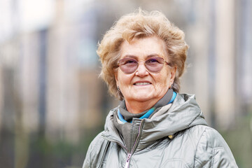Headshot of elderly woman smiling outside in urban scene. Mature woman looks friendly wearing glasses and jacket. Longevity and active senior living concept