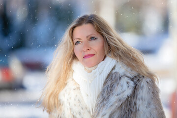 Beautiful adult blonde woman in a fur coat looks at the falling snow.