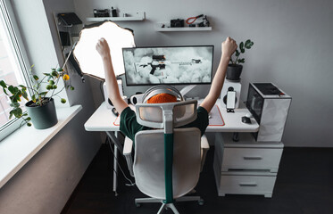 Teen girl celebrates gaming victory raising arms in excitement. Stylish gaming desk setup with headset, monitor and light panel in modern room. Joyful moment of success and digital lifestyle.