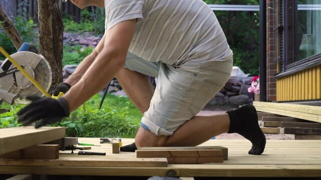 Man working on wooden deck construction outdoors using a circular saw, building a patio in backyard on a sunny day.