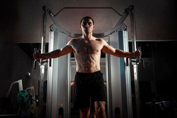 Fototapeta premium Training young man in a dark gym at night on a simulator for pumping the muscles of the biceps and triceps. Rendezvous of hands along with heaviness. The athlete is swinging.