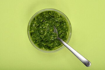 A glass bowl of vibrant green wild garlic (Allium ursinum) pesto with a silver spoon is presented on a solid light green background from a top-down perspective