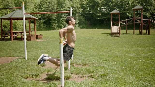 A young muscular man performs dips exercises on parallel bars outdoors. He completes a set of dips, demonstrating fitness and strength.