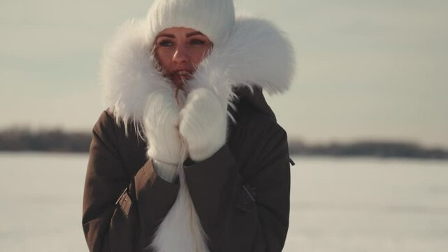 Winter-clad woman standing against snowy backdrop, bundled in white knitted accessories and fur-trimmed parka, battling cold with determined posture