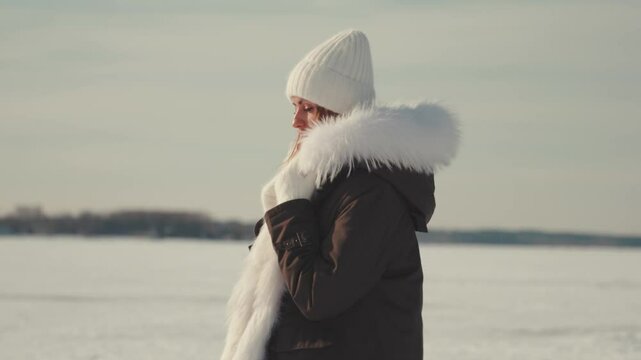 Fashion model wearing white woolen hat, fur-trimmed parka, posing in pristine snowy landscape, showcasing winter elegance against soft, white background