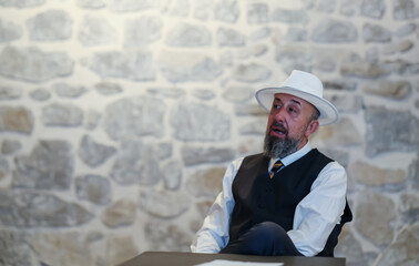 Elderly man in a white hat and vest gesturing expressively while sitting at a table, portraying a...