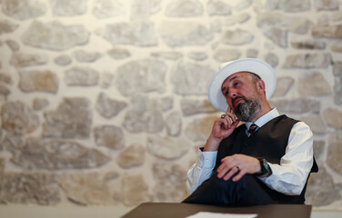 Elderly man in a white hat and vest gesturing expressively while sitting at a table, portraying a...