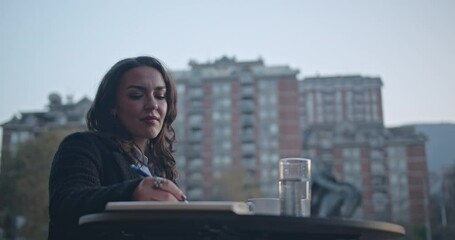 Young woman sitting at a table outside, writing in her notebook and drinking a coffee in an urban setting.