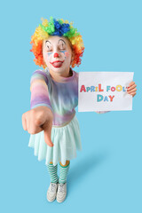 Funny little girl in clown wig with card pointing at viewer on blue background. April Fools' Day celebration