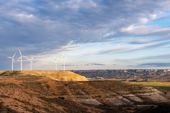Long row of wind turbines cutting across arid land in Zaragoza. Warm terrain, vivid sky and renewable energy structures in contrast with the natural topography.