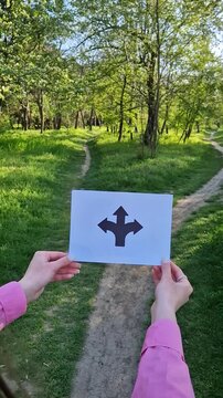 Woman hands holding a paper with arrows pointing left, right, and straight directions white standing in front of a crossroad. Forked path symbol for decision making and life choices concept