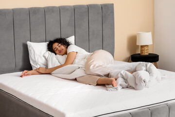 Young African-American woman with blanket lying on soft mattress in bedroom