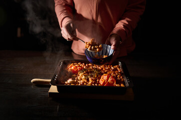 Person serving a hearty tray of baked beans and tomatoes into a bowl, set under warm, rustic...